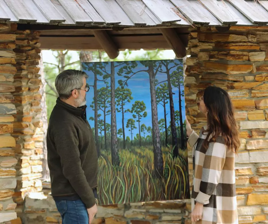 A man and woman standing under a wooden-roofed stone pavilion in Kisatchie National Forest, admiring a vibrant painting of tall pine trees and grasslands.