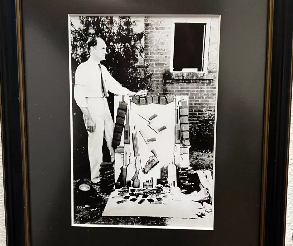 Historic black-and-white photo showing a lawman standing beside confiscated weapons and ammunition linked to Bonnie and Clyde.
