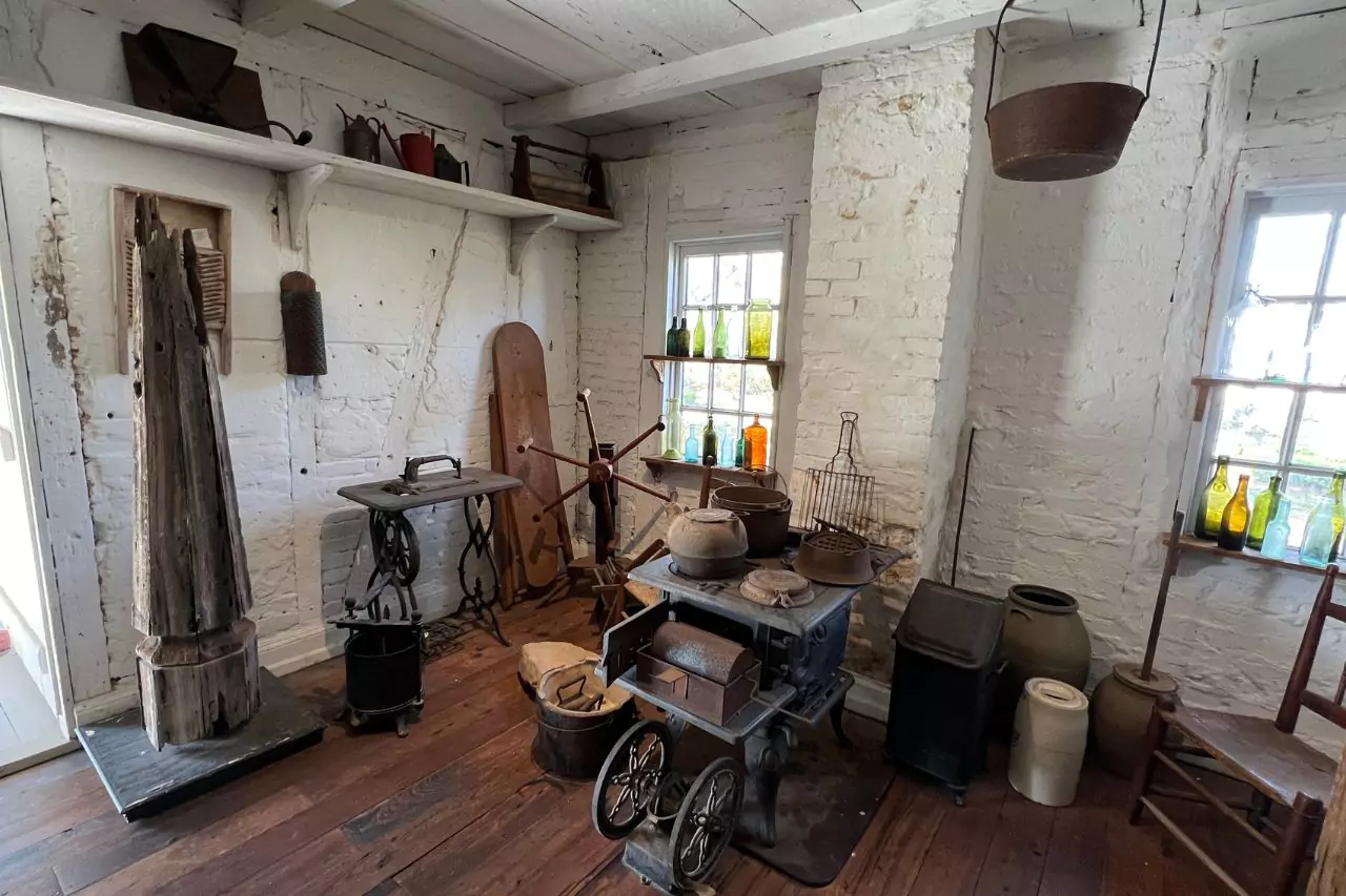 Historic Kitchen Tools and Antique Stove Display of vintage cooking tools, cast iron stove, colorful glass bottles, butter churn, and kitchenware in rustic white brick room at Lafayette Museum.