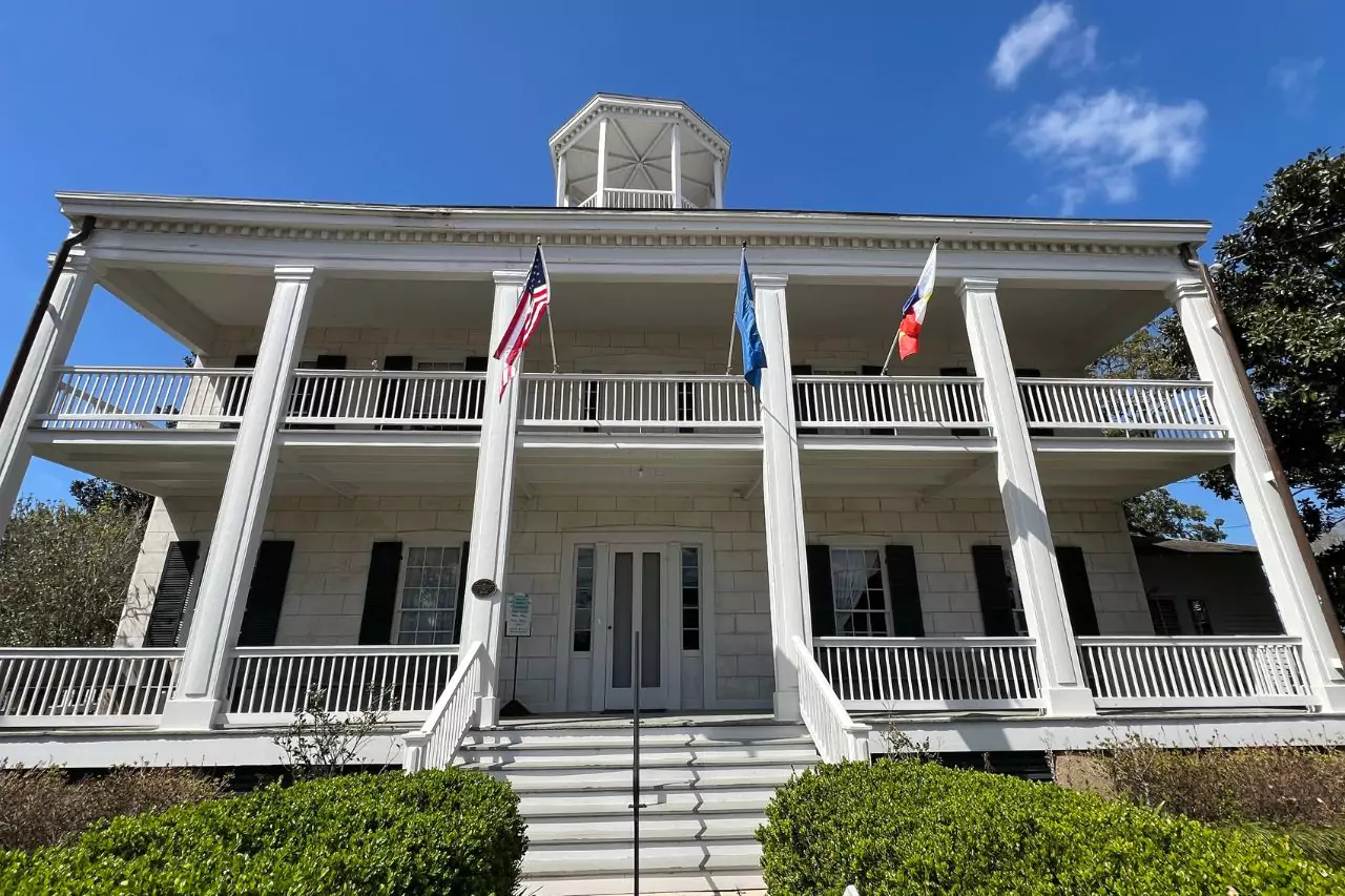 Front of Lafayette Museum - Alexandre Mouton House Historic Lafayette Museum front exterior with grand white columns, American, Louisiana, and French flags, two-story Greek Revival architecture, and manicured landscaping under a clear blue sky.