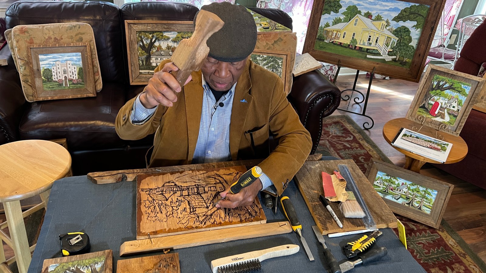 Cypress Wood Carver Henry Watson holds up a worn mallet while working on a detailed wood carving at his studio in New Roads, surrounded by framed pieces.