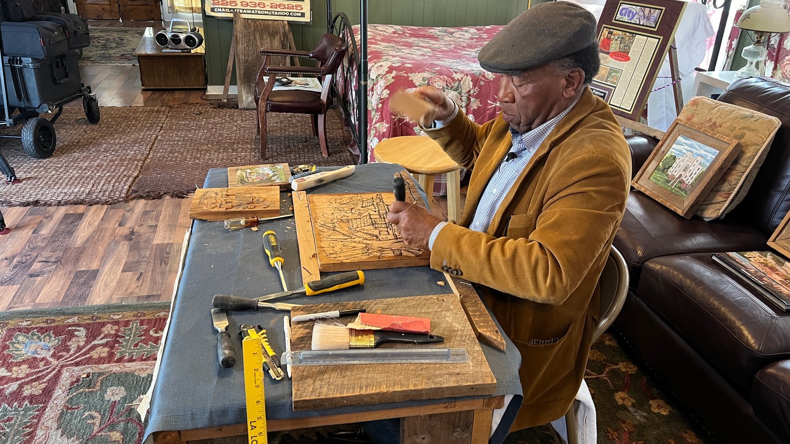 Henry Watson sits at his workbench mid-carving, surrounded by tools and framed cypress wood artworks inside his New Roads studio.