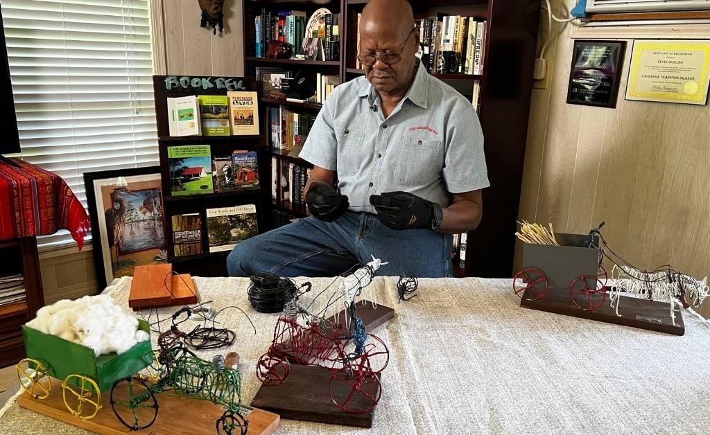 Elvin Shields sits in his home studio surrounded by handcrafted wire toys, including miniature wagons, animals, and cotton carts, with awards and books displayed in the background.