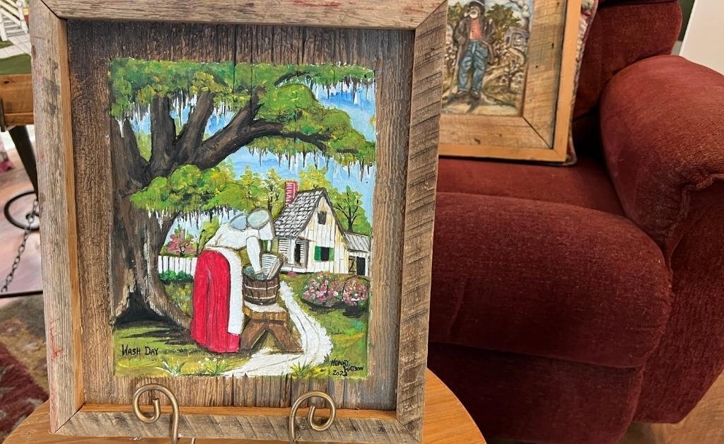 Framed wood relief titled “Wash Day” by Henry Watson, depicting a woman doing laundry under a mossy oak tree beside a Creole-style Louisiana cottage.
