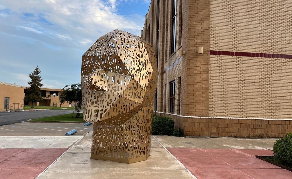 A golden metal head-shaped sculpture titled Here We Go by artist Beth Nybeck, located at the University of Louisiana Monroe, featuring cut-out letters formed from handwritten reflections by ULM students and community members.