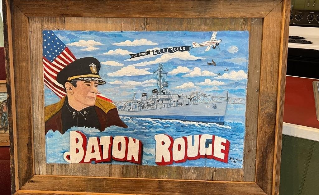 Framed painting depicting a Navy officer, the USS Kidd, and “Baton Rouge” in bold letters, honoring local history and service.
