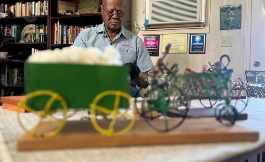 Elvin Shields works on wire sculptures at a table inside his cabin, with shelves of books and plaques on the wall behind him. A cotton-filled wagon and green tractor sit in the foreground.