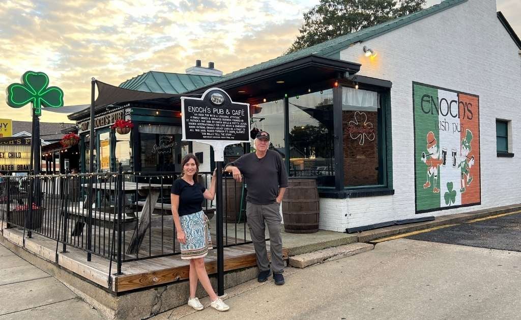 Karen LeBlanc stands beside Enoch Doyle Jeter in front of Enoch’s Pub & Café in Monroe, Louisiana, under the NELA Music Trail marker dedicated to the historic music venue. The Irish-themed building is adorned with a mural of dancing leprechauns and musical notes.