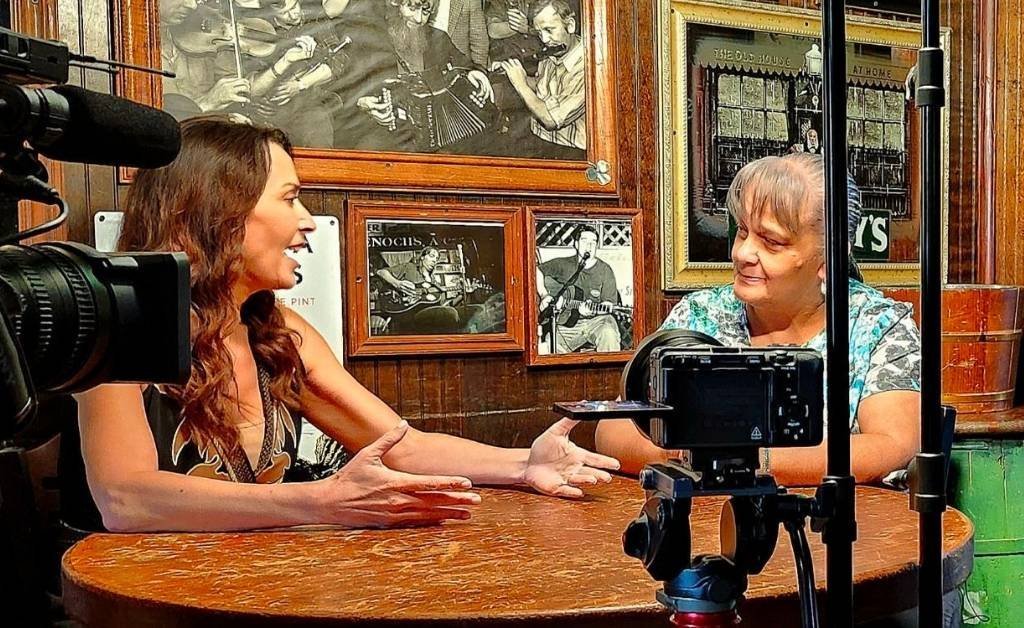 Karen LeBlanc with Brenda Roy, niece of Ivory Joe Hunter, seated at a wooden table in a music-themed venue decorated with framed historical photos and captured on video.