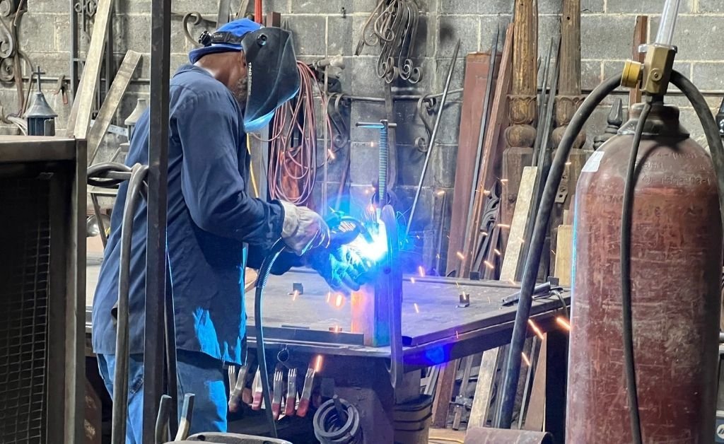 Master blacksmith Darryl Reeves welding inside his New Orleans 7th Ward workshop, sparks glowing as he works on an iron piece.