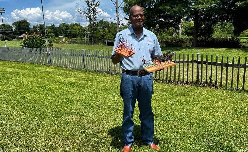 Elvin Shields stands on the grounds of Oakland Plantation holding two wire toy sculptures, with a wooden fence and trees in the background.