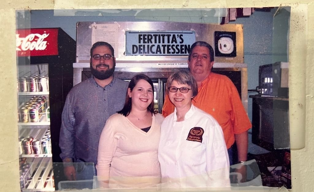 Family and staff members inside Fertitta's Delicatessen in Shreveport, standing together in front of the deli counter with the vintage Fertitta's sign overhead.