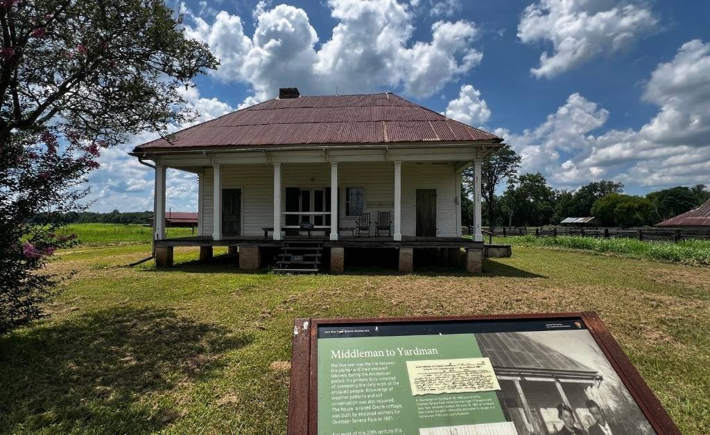 A restored white Creole cottage at Oakland Plantation under a blue sky, with an informational panel in the foreground describing the role of overseers and yardmen.