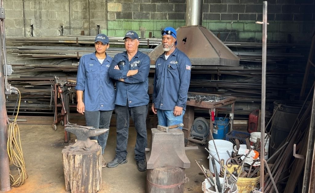 Darryl Reeves with apprentice Karina Roca and a fellow craftsman inside his New Orleans forge, standing by anvils and the coal-fired hearth.