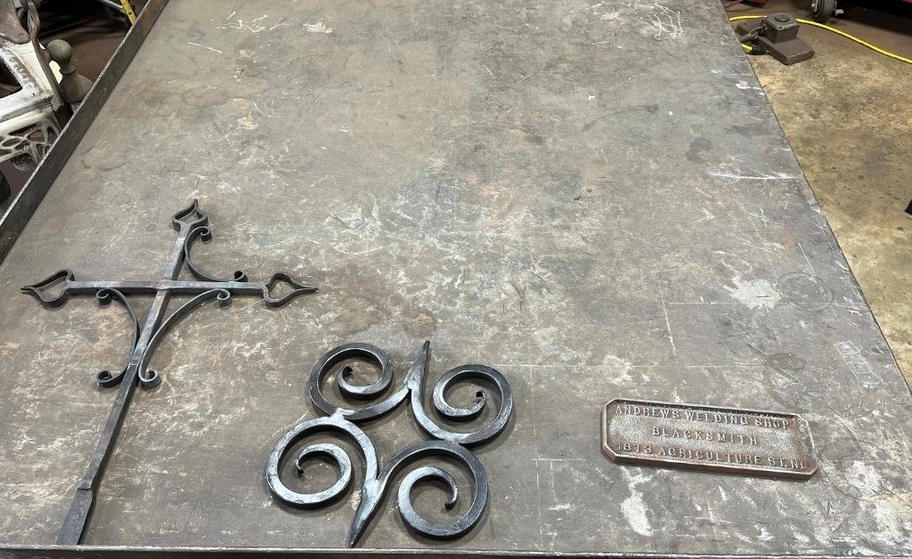Hand-forged iron designs and a shop plaque from Andrew’s Welding & Blacksmith Shop displayed on a worktable in Darryl Reeves’ forge.