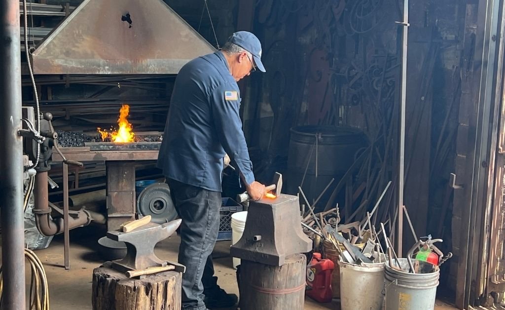 Master blacksmith Darryl Reeves shaping heated iron on an anvil inside his New Orleans 7th Ward forge, with flames glowing in the background.