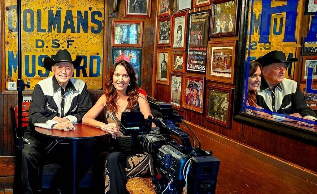 Karen LeBlanc sits with Gene Stewart at Enoch’s Irish Pub, surrounded by signed memorabilia, vintage photos, and a film crew documenting their conversation.