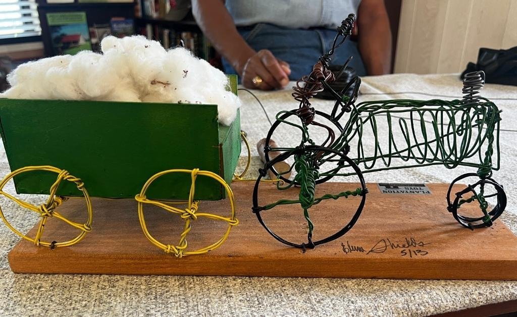 Close-up of a handcrafted plantation toy by Elvin Shields, featuring a green wagon filled with cotton and a wire-formed tractor, mounted on a wooden base and signed by the artist.