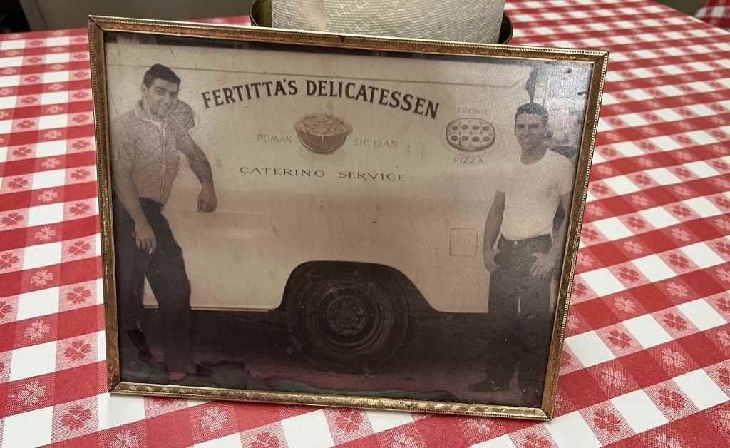 Framed vintage photo of two men standing beside a Fertitta's Delicatessen catering truck, displayed on a red-checkered table inside the deli.