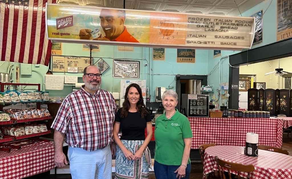 Karen LeBlanc stands inside Fertitta's Deli in Shreveport with Reverend Patrick Fertitta and Agatha Fertitta McCall, surrounded by vintage signage and red-checkered tables.