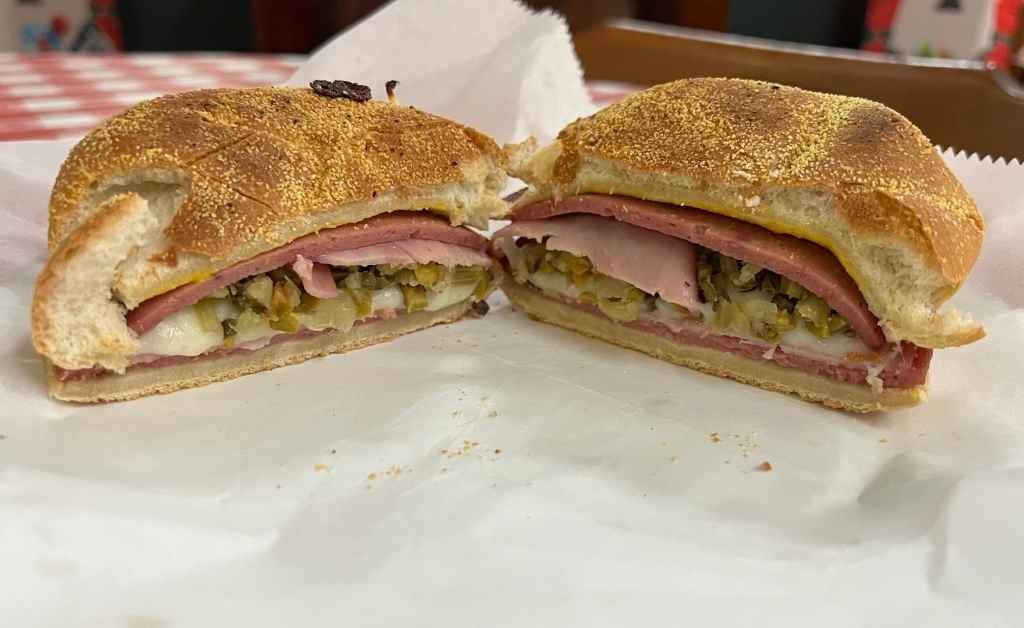 Close-up of the Muffy sandwich at Fertitta's Deli, showing layers of ham, salami, mozzarella cheese, and chopped olive salad on cornmeal-dusted bread.