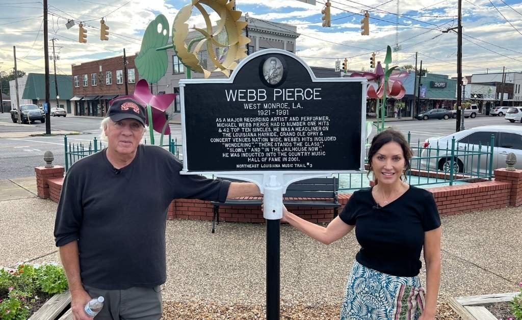 Karen LeBlanc and Enoch Doyle Jeter stand beside the Northeast Louisiana Music Trail marker honoring country star Webb Pierce in downtown West Monroe. The marker is placed near a decorative fence with downtown shops and traffic lights visible in the background.