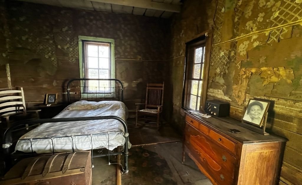 Interior of a preserved one-room cabin at Oakland Plantation, showing a metal-frame bed, wooden dresser, and weathered walls with period furnishings.