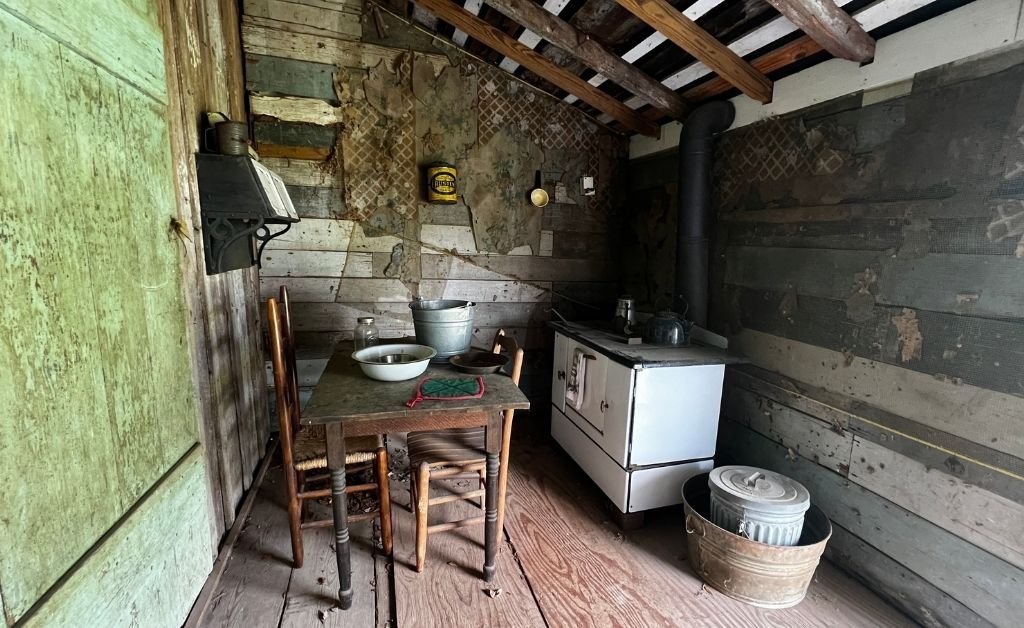 Interior of a historic plantation cabin kitchen with exposed beams, a wood-burning stove, rustic furniture, and weathered walls reflecting early 20th-century life.