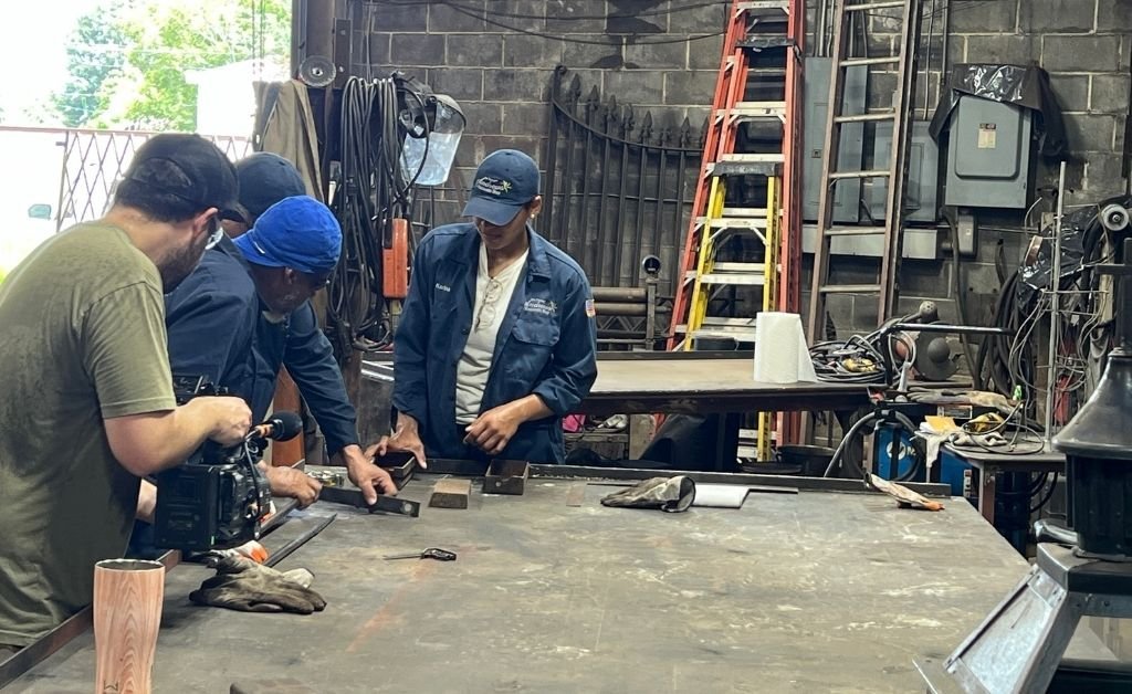 Darryl Reeves and apprentice Karina Roca working with a team inside the 7th Ward forge, examining pieces of hand-forged iron on a workshop table.