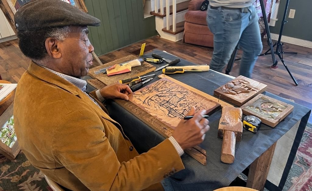 Henry Watson sits at a table in his studio, sketching a new cypress wood carving surrounded by his chisels, mallet, and finished relief artworks.