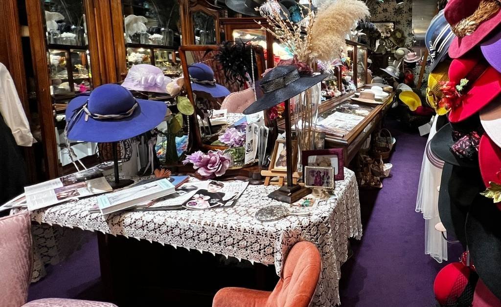 A lace-covered vanity table at Yvonne LaFleur’s boutique displaying custom hats, perfume bottles, silk flowers, and framed photos, surrounded by antique mirrors and jewelry cases.