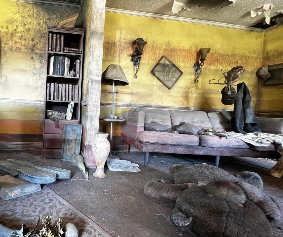 Room with water-stained yellow walls, a sofa covered in debris, scattered belongings, and damaged furniture preserved to show the aftermath of Hurricane Katrina’s London Avenue Canal floodwall breach.