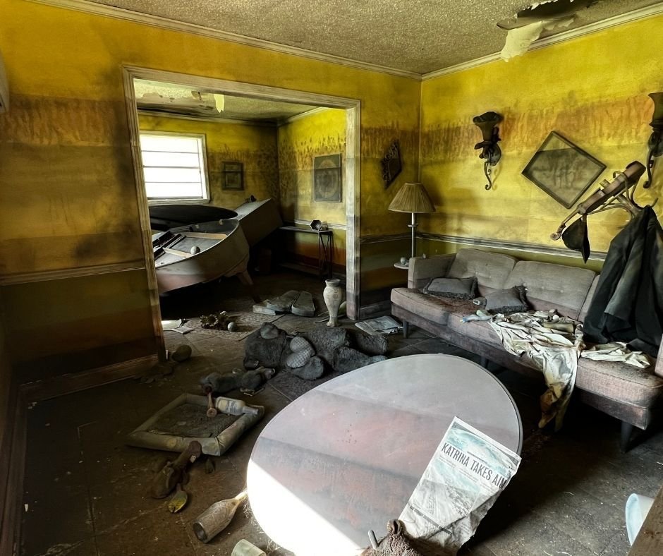 Interior of the Flooded House Museum in New Orleans, showing water-stained walls, overturned furniture, scattered belongings, and a newspaper with the headline “Katrina Takes Aim,” preserved to depict the aftermath of Hurricane Katrina.