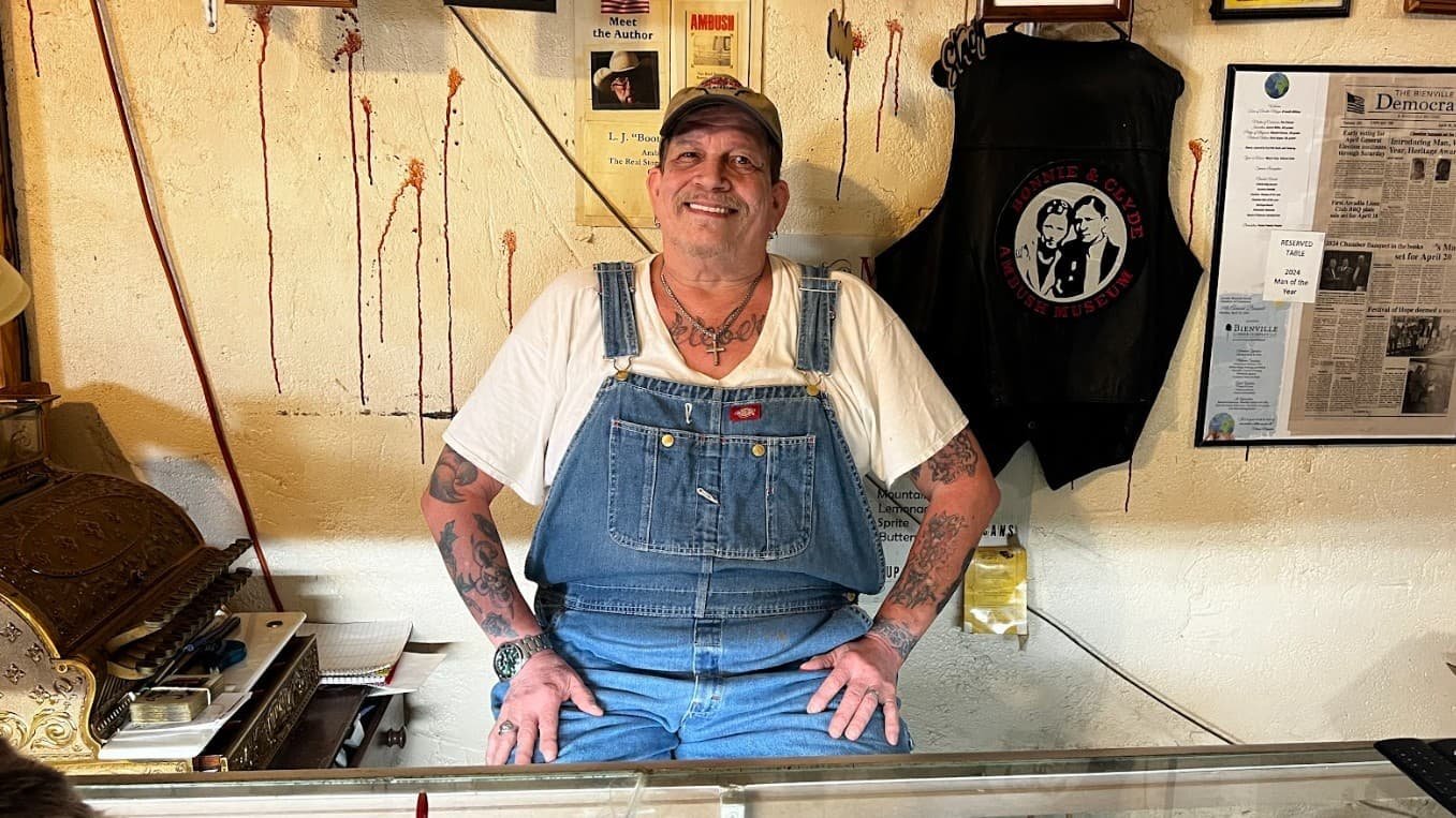 Perry Carver, owner of the Bonnie and Clyde Ambush Museum in Gibsland, Louisiana, smiling behind the front counter surrounded by memorabilia.