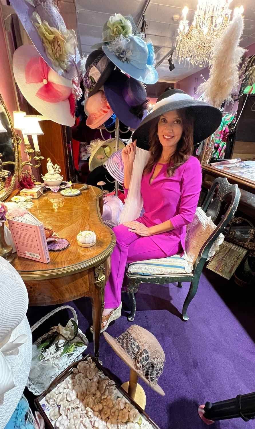 Karen LeBlanc wearing a wide-brimmed hat inside Yvonne LaFleur’s boutique, seated at a vintage vanity surrounded by custom hats.