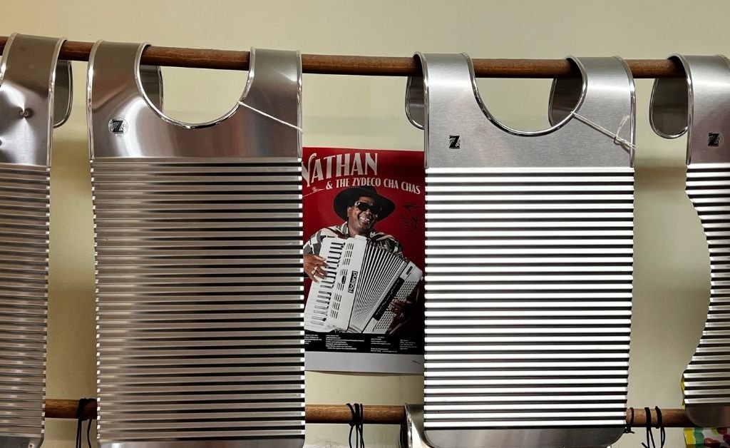 Close-up of three handcrafted stainless steel Zydeco rubboards made by Tee Don Landry, hanging on a wooden rod with a Zydeco-themed poster in the background.