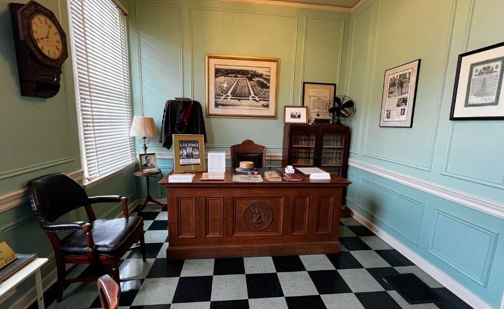 Historic office room inside the Old Governor's Mansion, featuring a wooden desk with memorabilia, framed photos and documents on pastel blue paneled walls, black-and-white checkered flooring, and period furnishings including chairs and a vintage clock.