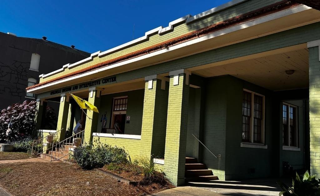 Wide view of the green brick Opelousas Museum and Interpretive Center, showing its entrance, columns, and yellow flag.