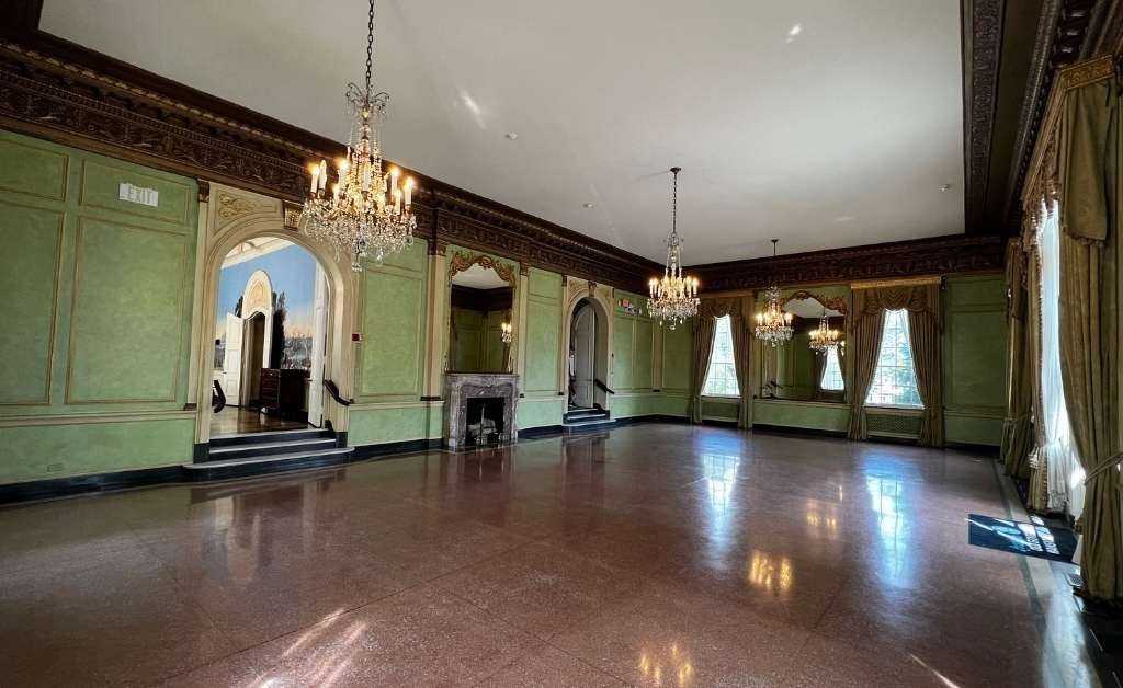 Spacious ballroom inside the Old Governor's Mansion featuring green walls, crystal chandeliers, large arched windows with drapes, and polished flooring. Decorative crown molding and a marble fireplace add to the room's elegance.
