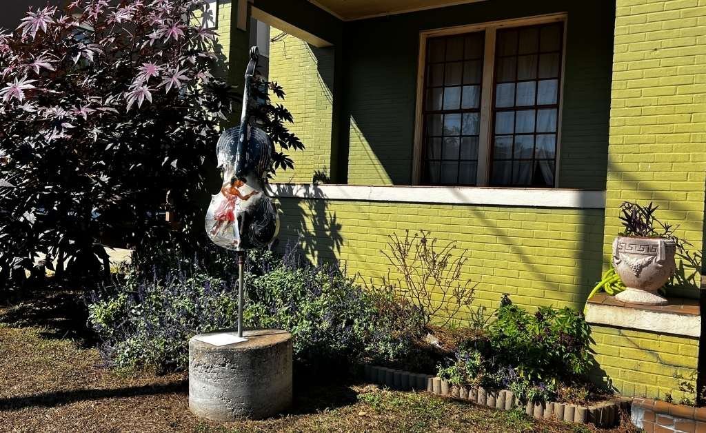 A painted sculpture of a cello mounted on a pedestal in a garden area outside a green brick building.