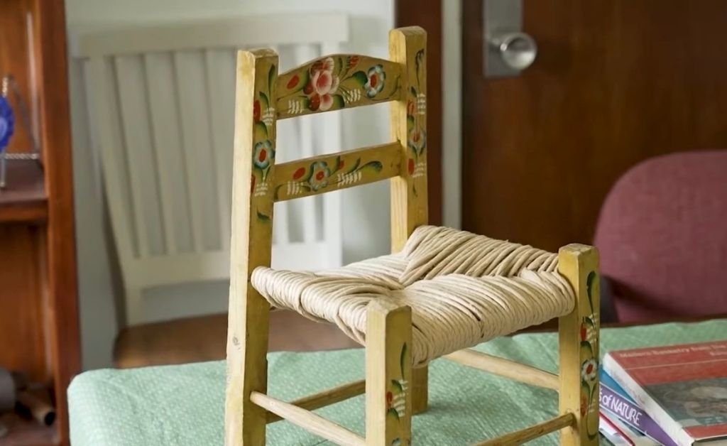A hand-woven cane chair with painted floral details sits on a table beside books, showcasing traditional Louisiana French craftsmanship taught by Mavis Fruge.