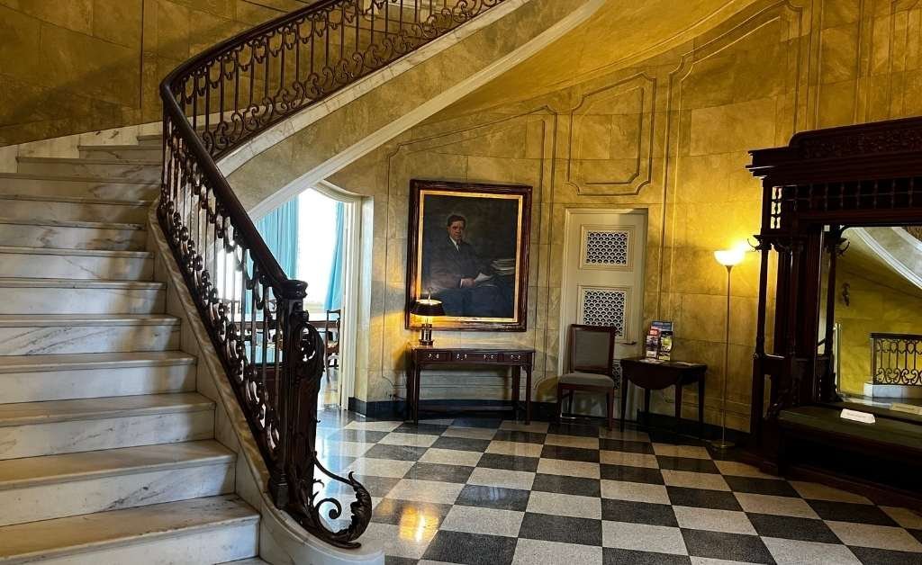 Grand staircase inside the Old Governor's Mansion featuring white marble steps, ornate wrought-iron railing, black-and-white checkered floor, and a portrait of Huey P. Long hanging on a gold-toned wall near period furniture and vintage lighting.