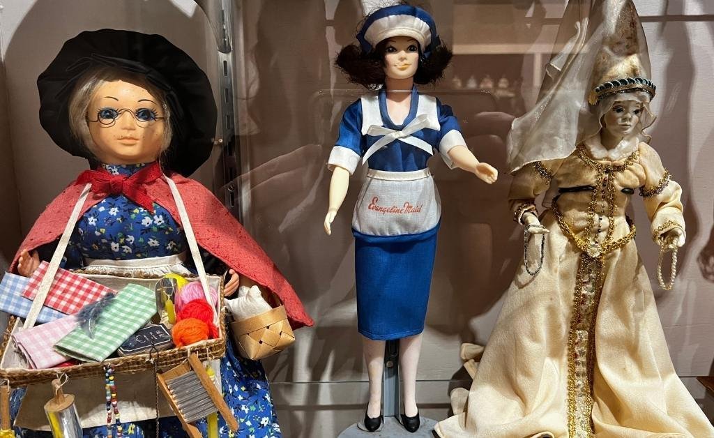 Three collectible dolls displayed at the Opelousas Museum, representing different cultural roles: a fabric vendor, an Evangeline Maid bakery worker, and a robed ceremonial figure.