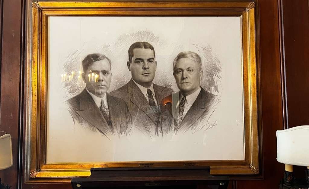 A framed black-and-white portrait of three men in suits, drawn in a realistic style, hanging on a wood-paneled wall between two wall lamps.