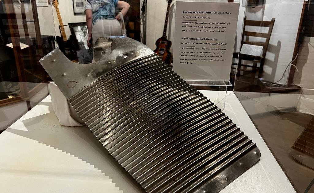 Metal washboard-style vest, known as a frottoir, used in Zydeco music, displayed at the Opelousas Museum with informational placards and instruments in the background.