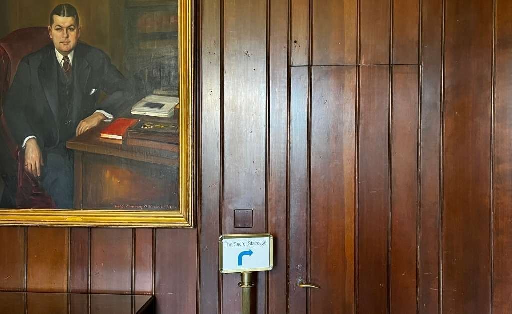 A portrait of Huey P. Long hanging beside a wood-paneled wall in the Old Governor's Mansion library. A small sign labeled "The Secret Staircase" points to a hidden door built into the paneling, which leads to a private stairway.