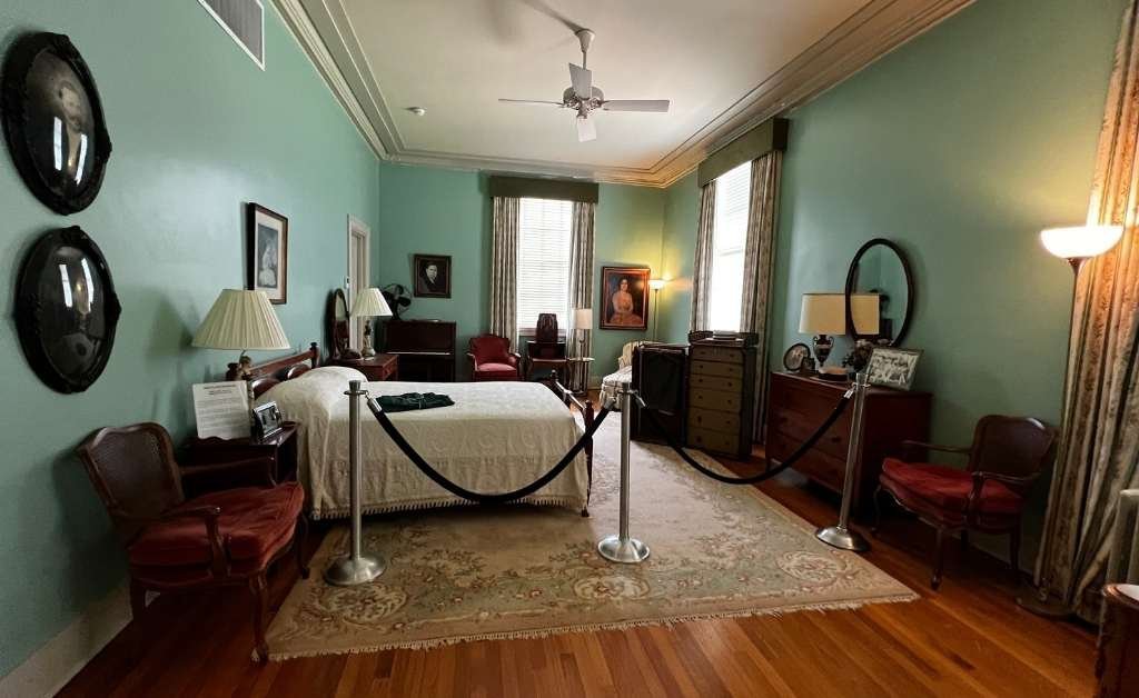 Interior view of Huey P. Long's bedroom at the Old Governor's Mansion, featuring vintage furniture including a bed, dressers, nightstands, and chairs. The room has green walls, hardwood floors, two tall windows with curtains, and framed portraits. A roped barrier surrounds the bed for preservation.