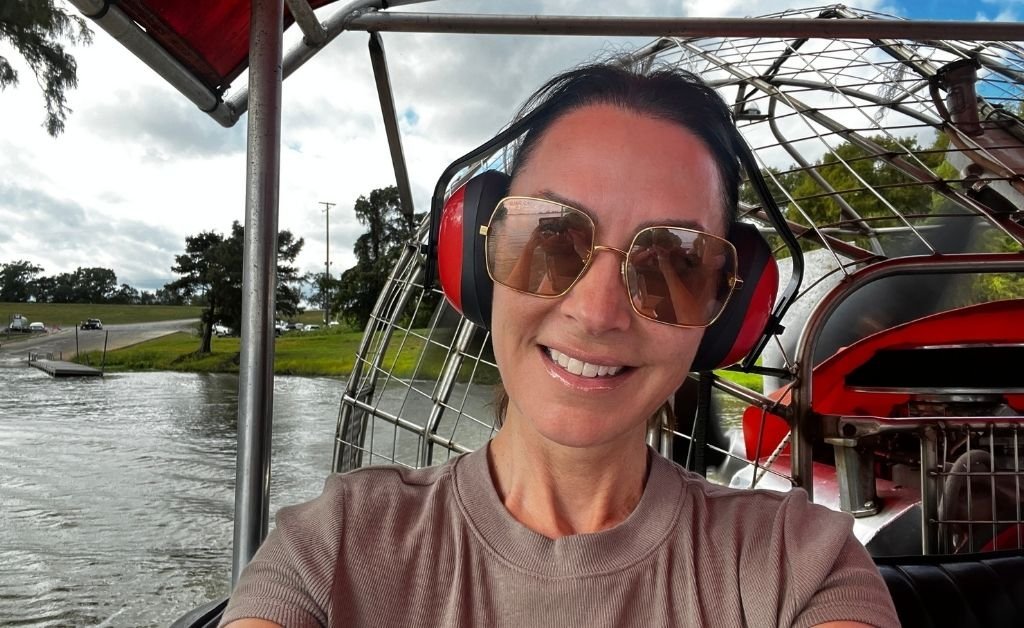 Karen LeBlanc wearing protective earmuffs and sunglasses, smiling on an airboat at Atchafalaya Basin Landing, with swamp water and a boat ramp in the background.