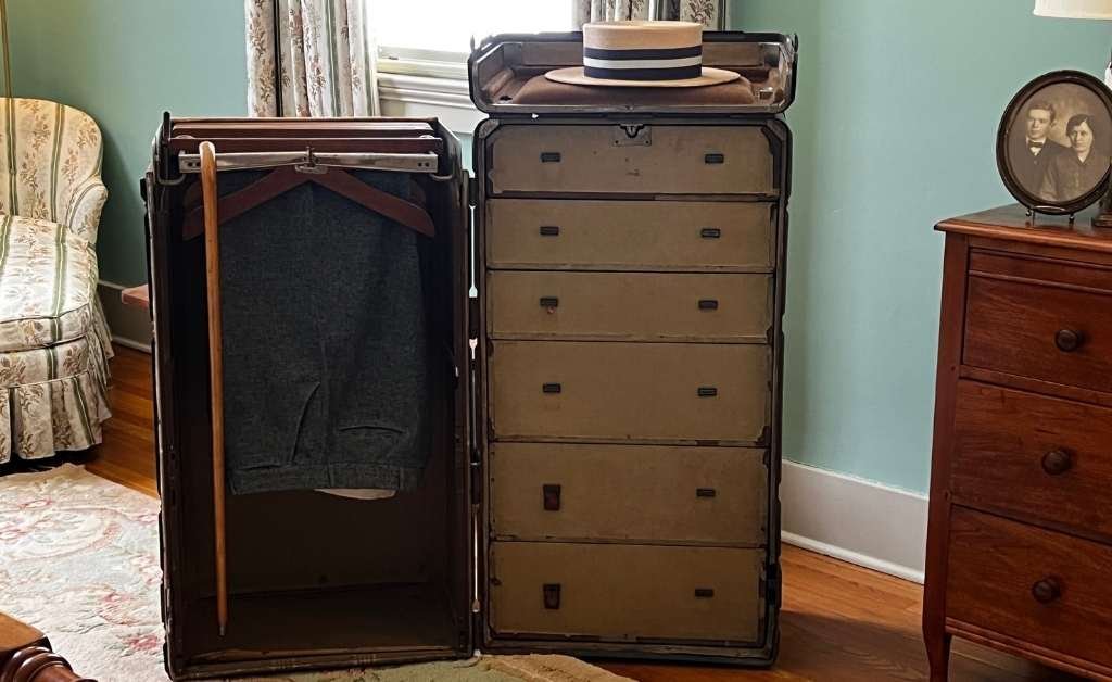 An open vintage wardrobe trunk in Huey P. Long's bedroom, displaying a gray suit, a wooden cane, and a straw boater hat, with a framed family photo and antique furniture nearby.