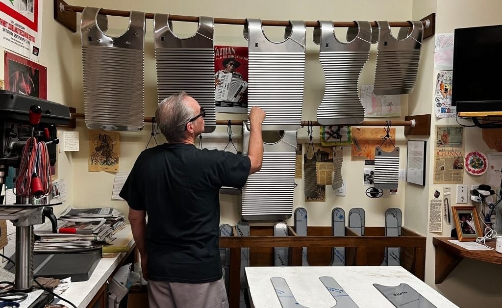 Tee Don Landry stands in his workshop, surrounded by stainless steel rubboards hanging on wooden rods. The room is decorated with music memorabilia, press clippings, and rubboard patterns on the table.